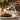 Steak with vegetables being drizzled with sauce, set on a table with a glass of wine and flowers.
