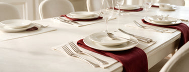 A styled dining table setting with white plates, silver cutlery, clear wine glasses, and deep burgundy napkins and placemats on a white tablecloth.